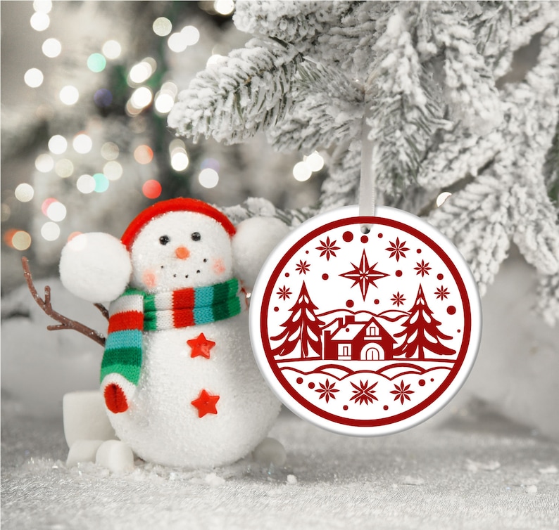 May include: A white ceramic Christmas ornament with a red design of a house, two pine trees, and a star in a snowy landscape.