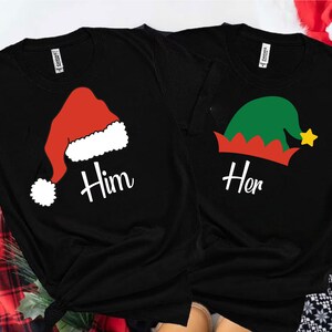 May include: Two black t-shirts with a Christmas theme. One shirt features a red Santa hat with white trim and the word "Him" printed on it. The other shirt has a green elf hat with red trim and the word "Her" printed on it.