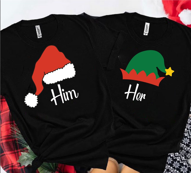 May include: Two black t-shirts with a Christmas theme. One shirt features a red Santa hat with white trim and the word "Him" printed on it. The other shirt has a green elf hat with red trim and the word "Her" printed on it.