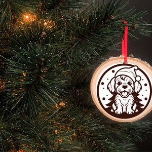 May include: A round wooden Christmas ornament with a white silhouette of a dog wearing a Santa hat. The ornament is hanging from a red ribbon on a green Christmas tree.