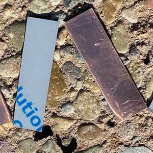 May include: Five rectangular strips of material, two are copper colored and three are gray. The copper strips have a slightly rough texture. The gray strips have a smooth, slightly shiny surface. The strips are arranged in a V shape.