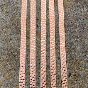 May include: Five copper strips with a hammered texture, arranged vertically against a mottled gray background. The strips have a rectangular shape and a warm, reddish-brown color. The hammered texture creates a pattern of small, irregular indentations.