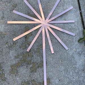 May include: A copper-colored metal starburst design with 12 points radiating out from a central point. The starburst is laid flat on a gray concrete surface.