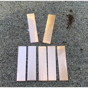 May include: Seven rectangular copper metal pieces are arranged on a textured gray surface. Two pieces are positioned above five others. The copper has a shiny, reflective surface, with variations in color from light to dark.