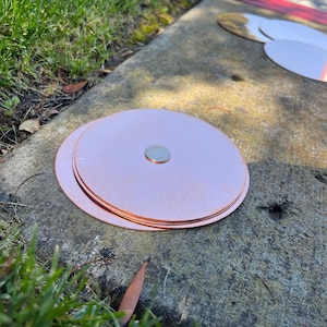 May include: A stack of copper-colored, circular metal discs with a silver coin on top. The discs are on a concrete surface, with green grass and a red-painted edge in the background. The discs appear to be new.