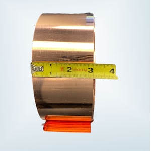 May include: A roll of copper foil, approximately 3 inches in diameter, is measured with a yellow Stanley tape measure. The copper has a shiny, metallic finish. The tape measure is positioned horizontally across the roll.