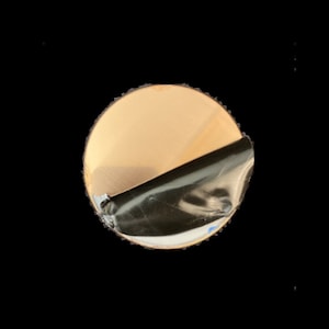 May include: A round wooden coaster with a brown wood grain finish. The coaster has a black backing with a peel-off adhesive.