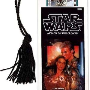 May include: A black and white bookmark featuring a movie poster for Star Wars Episode II: Attack of the Clones. The poster features a red and orange background with images of Anakin Skywalker, Padmé Amidala, and Obi-Wan Kenobi. The bookmark has a black tassel.
