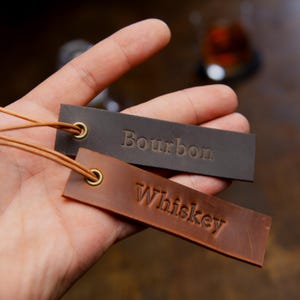 May include: Two rectangular leather tags, one dark brown and one light brown, are held in a hand. The dark brown tag reads "Bourbon" and the light brown tag reads "Whiskey". Both tags have a gold grommet and a thin leather cord.