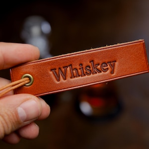 Könnte beinhalten: Ein rechteckiges, hellbraunes Lederetikett mit dem eingeprägten Wort Whiskey in der Mitte. Das Etikett verfügt über eine messingfarbene Metallöse auf der linken Seite, durch die ein dünnes Lederband zur Befestigung gezogen ist.