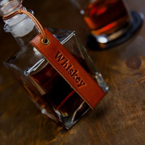 Könnte beinhalten: Eine klare Whisky-Karaffe aus Glas mit einem Lederanhänger mit der Aufschrift "Whiskey". Die Karaffe ist mit bernsteinfarbener Flüssigkeit gefüllt und hat einen Glasverschluss. Ein Glas Whisky befindet sich im Hintergrund.