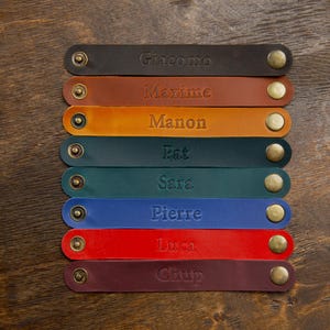 May include: A collection of personalized leather cable organizers in various colors, including brown, orange, green, blue, red, and burgundy. Each organizer is embossed with a name and secured with brass hardware.