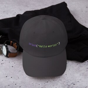 May include: A dark gray baseball cap with the code "print("Hello world!")" embroidered in purple and green.