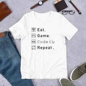 May include: White t-shirt with the text "Eat. Game. Code (); Repeat." in black. The shirt features icons of a pizza slice, a game controller, code brackets, and a repeat symbol. The shirt is paired with blue jeans and casual shoes.