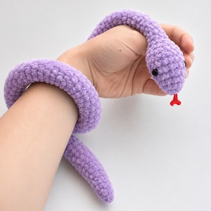 Puede incluir: Una serpiente de ganchillo color lavanda hecha de hilo suave y afelpado. La serpiente presenta una cabeza redondeada con dos pequeños ojos de cuentas negras y una lengua bífida de color rojo brillante que sobresale de su boca.