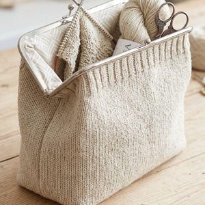 Könnte beinhalten: Eine beige Stricktasche mit silbernem Verschluss, geöffnet, um Stricknadeln, Garn und eine Schere zu zeigen. Die Tasche ist rechteckig und steht auf einem Holztisch. Das Innere der Tasche hat ein gemustertes Futter.