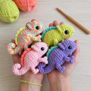 May include: Four small, handmade crochet chameleon toys made from soft, plush yarn. The chameleons feature textured stitches, curled tails, and dark plastic safety eyes. They are coloured in variegated pastel, solid lime green, solid pink, and solid purple.
