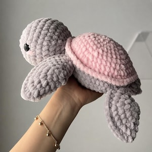 Peut inclure: Une peluche en forme de tortue fabriquée à partir de fil gris épais et doux, dotée d'une carapace arrondie d'une couleur rose clair contrastante. La tortue présente des yeux en plastique noir lisse et une surface crochetée texturée.