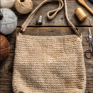 May include: A beige crocheted shoulder bag with a long strap, set on a wooden surface. The bag is surrounded by crochet hooks, yarn balls, a thimble, and scissors, suggesting a crafting theme. The bag's dimensions are approximately 12 inches wide and 14 inches tall.