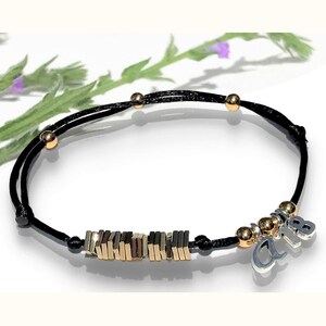 Puede incluir: Pulsera de cordón negro con cuentas y detalles rectangulares dorados. Un dije plateado con las letras "AB" cuelga de la pulsera. La pulsera se exhibe sobre una superficie blanca con follaje verde en el fondo.