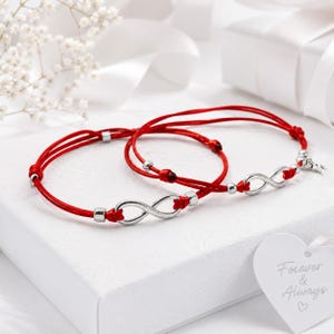 May include: Two red cord bracelets featuring silver-toned infinity symbols and small metallic beads. Each bracelet has a sliding knot closure. A white heart-shaped tag nearby displays the text Forever & Always with a small heart icon.
