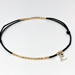 Puede incluir: Un brazalete de cordón negro con una barra dorada y un dije de plata en forma de la letra L.