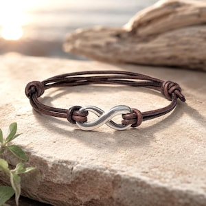 May include: A bracelet featuring a silver-toned infinity symbol charm centered on multiple strands of dark brown leather cord. The leather cords are secured with knots on either side of the metal charm, creating a layered and adjustable design.