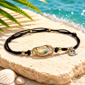 May include: A black cord bracelet featuring a central gold-toned flip-flop charm inlaid with iridescent shell material and accented with small clear crystals. The bracelet includes gold-toned metal beads and a small silver-toned wave-shaped charm.