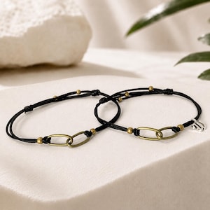 May include: Two black cord bracelets featuring small metallic gold-toned beads and two interlocking oval brass-colored links. One bracelet includes a small silver-toned charm shaped like the letter M. The bracelets have a thin, adjustable design.