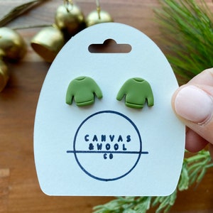 Könnte beinhalten: Olivgrüne Ohrstecker in Form eines Pullovers auf einer weißen Karte mit dem Text "Canvas & Wool Co." Die Ohrringe sind klein und detailliert, mit einem Miniatur-Pullover-Design. Die Karte wird von einer Person gehalten.