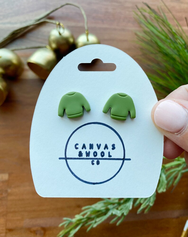 K&ouml;nnte beinhalten: Olivgr&uuml;ne Ohrstecker in Form eines Pullovers auf einer wei&szlig;en Karte mit dem Text "Canvas & Wool Co." Die Ohrringe sind klein und detailliert, mit einem Miniatur-Pullover-Design. Die Karte wird von einer Person gehalten.