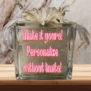 May include: A clear glass block with a white ribbon and gold accents. The block has the text "Make it yours! Personalize without limits!" in pink lettering.