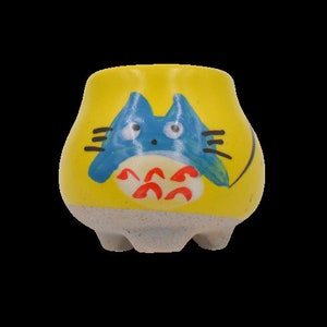 Puede incluir: Una pequeña maceta de cerámica amarilla con un personaje de dibujos animados azul y rojo pintado en la parte delantera. El personaje tiene ojos grandes y un cuerpo redondo.