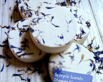 May include: Four white shower steamers with blue and brown flower petals on a white wooden surface. The text "Scorpio Scents Shower Steamers" is on a blue label.