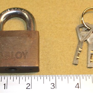 Puede incluir: Un candado de color bronce con la palabra "ABLOY" grabada en la parte delantera. El candado tiene un grillete de metal curvado y está acompañado de tres llaves en un anillo. La imagen está tomada sobre un fondo marrón claro.