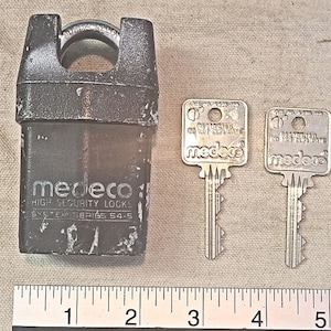 Puede incluir: Una cerradura de seguridad alta medeco negra con dos llaves plateadas. Las llaves están etiquetadas como "Llave restringida No duplicar" y "medeco".