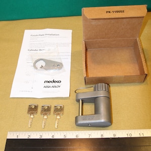 Può includere: Un lucchetto argentato con un foro per la chiave e una chiave argentata. Il lucchetto è etichettato "medeco ASSA ABLOY". Sono incluse tre chiavi dorate. Il lucchetto è in una scatola di cartone marrone con l'etichetta "PK-110052".