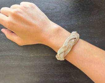 May include: A rose gold-toned cuff bracelet featuring a braided design. The surface of the bracelet is densely encrusted with numerous small, clear, reflective stones that catch the light, creating a textured and shimmering appearance across the band.