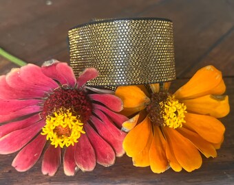 May include: A wide cuff bracelet featuring a dense, textured surface of shimmering gold-toned micro-crystals. The bracelet is positioned behind a vibrant pink zinnia flower with a yellow center and an orange zinnia flower with a yellow center.