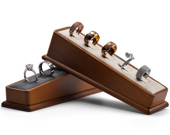 May include: A wooden ring display stand with five slots for rings. The stand is made of light brown wood and has a velvet lining. There are five rings on the stand, including a silver ring with a pink stone, a silver band, a gold band, a rose gold band, and a silver ring with a swirl design.