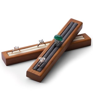 Op de afbeelding: Twee houten ringdisplay trays met een donkere afwerking. Eén tray heeft een lichtgekleurd interieur, met twee ringen met heldere stenen. De andere tray heeft een grijs interieur, met een ring met een groene steen en een ring met heldere stenen.
