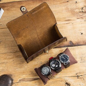 Peut inclure: Un porte-montres en cuir marron avec trois montres &agrave; l'int&eacute;rieur. Les montres ont des cadrans noirs et des bracelets argent&eacute;s.