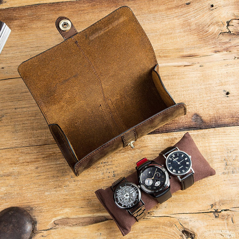 Peut inclure: Un porte-montres en cuir marron avec trois montres &agrave; l'int&eacute;rieur. Les montres ont des cadrans noirs et des bracelets argent&eacute;s.
