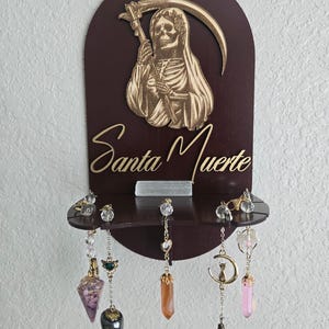 Pode incluir: Uma decoração de parede de madeira castanha escura com uma imagem cortada a laser de Santa Muerte segurando uma foice. O texto "Santa Muerte" está escrito em dourado. Abaixo, uma prateleira com brincos de cristal pendurados.