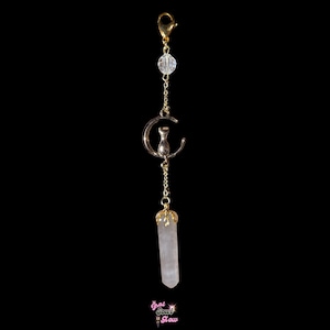 May include: A gold-colored charm with a cat-in-moon pendant, clear crystal beads, and a light pink crystal point. The charm is attached to a gold-colored clasp. The bottom of the charm has a light pink crystal point. The charm is against a black background.