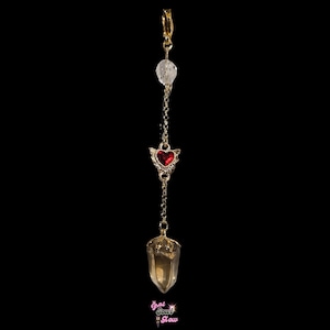 May include: A gold-colored pendulum with a clear crystal bead, a red heart-shaped charm with wings, and a pointed crystal at the bottom. The pendulum is suspended from a clip. The text "That Soul Glow" is at the bottom.