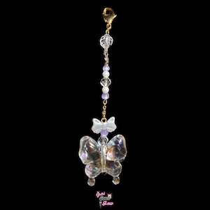 May include: A clear, iridescent butterfly charm with a gold-colored clasp and chain. The charm features a small white bow and is adorned with clear and purple beads. The words "that soul glow" are at the bottom.