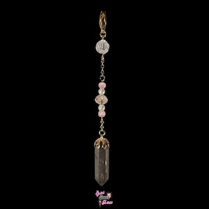 May include: A gold-colored charm with a clear crystal, pink and white beads, and a smoky quartz crystal pendant. The charm is suspended from a gold clasp. The bottom of the image has the text "That Soul Glow".