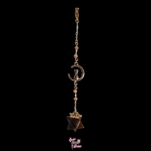 May include: A gold-colored charm featuring a cat on a crescent moon, with a star-shaped pendant. The charm is suspended by a chain with small, round, peach-colored beads. The background is black.
