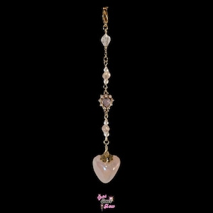 May include: A gold-colored hair accessory with a dangling design. The accessory features a chain with clear crystal beads, faux pearls, and a pink heart-shaped pendant. The bottom pendant is a larger, pearlescent pink heart.